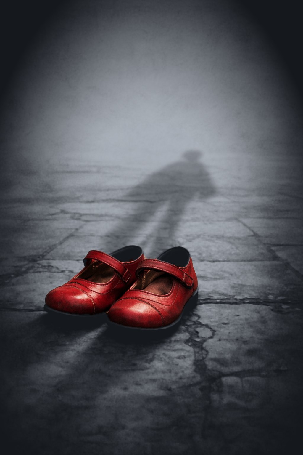 A pair of worn red children&rsquo;s shoes on grey stone, with the shadow of a child falling across the ground behind them